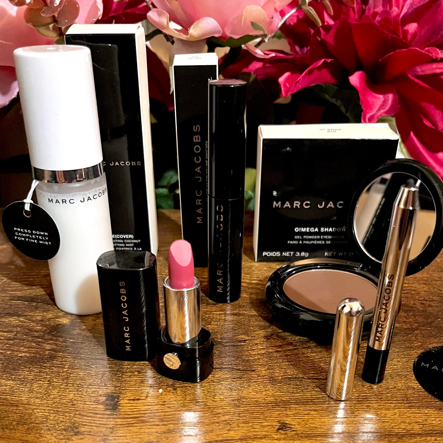 Elegant Marc Jacobs 6-piece luxury beauty gift collection featuring a glossy compact omega eyeshadow in the color “oh Boy”, A Limited Edition Signature Coconut Setting Spray, The Famous Award winning Highliner Gel Eye Crayon in the color “Blacquar” and an assortment of matching designer beauty cosmetics, complemented by a backdrop of lush pink, green & white florals.