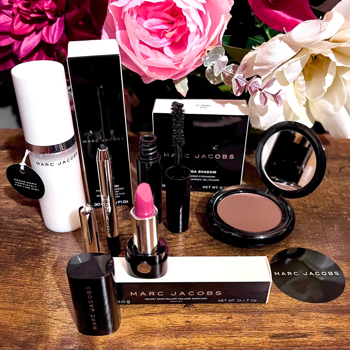 Elegant Marc Jacobs 6-piece luxury beauty gift collection featuring a glossy compact omega eyeshadow in the color “oh Boy”, A Limited Edition Signature Coconut Setting Spray, The Famous Award winning Highliner Gel Eye Crayon in the color “Blacquar” and an assortment of matching designer beauty cosmetics, complemented by a backdrop of lush pink, green & white florals.