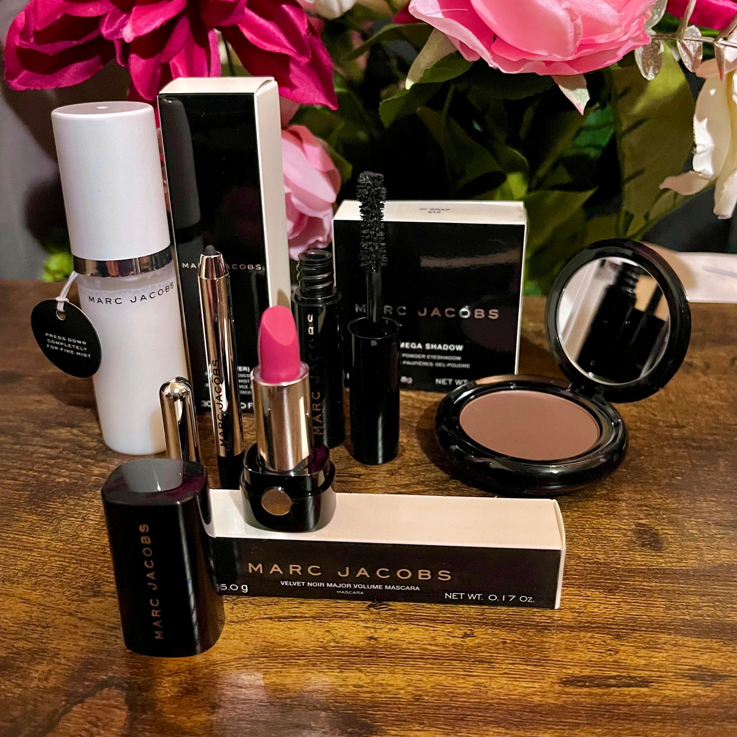 Elegant Marc Jacobs 6-piece luxury beauty gift collection featuring a glossy compact omega eyeshadow in the color “oh Boy”, A Limited Edition Signature Coconut Setting Spray, The Famous Award winning Highliner Gel Eye Crayon in the color “Blacquar” and an assortment of matching designer beauty cosmetics, complemented by a backdrop of lush pink, green & white florals.