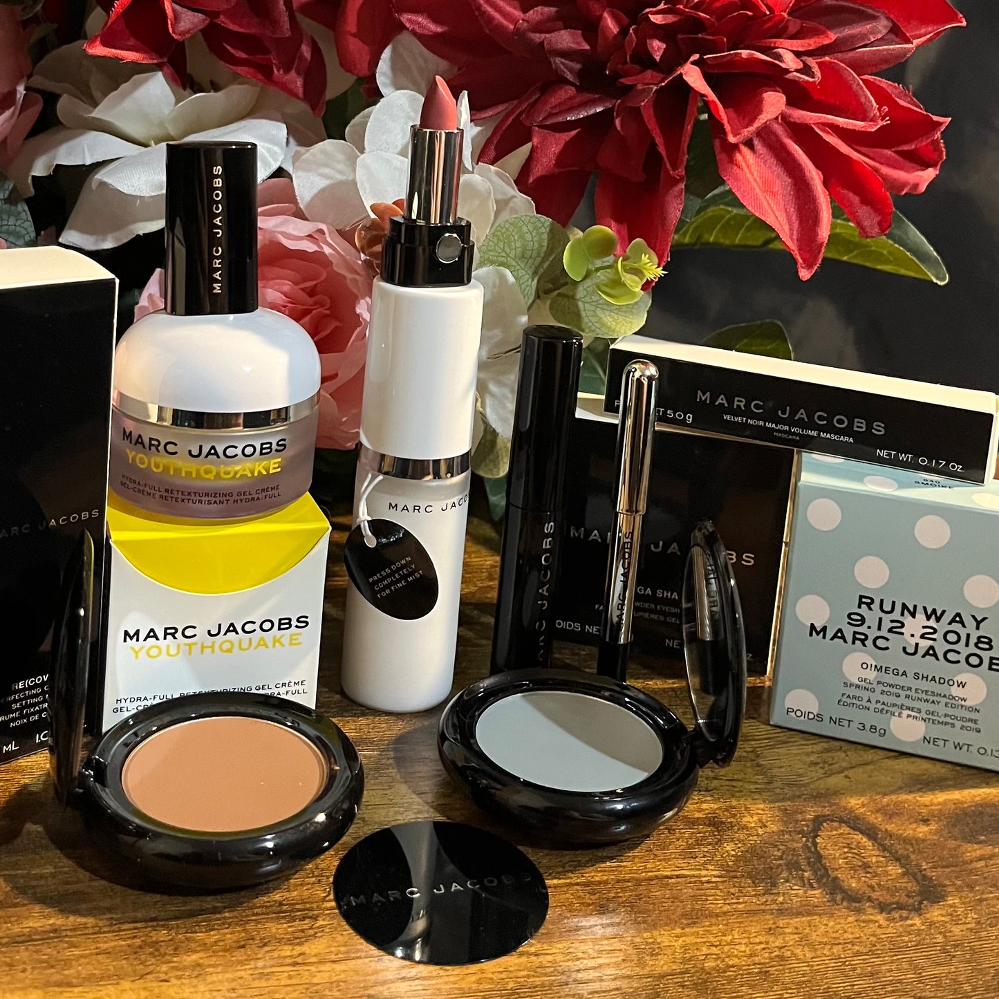 Elegant Marc Jacobs beauty collection featuring glossy compact powders, signature coconut setting spray, and assorted beauty cosmetics, complemented by a backdrop of lush pink & white florals.