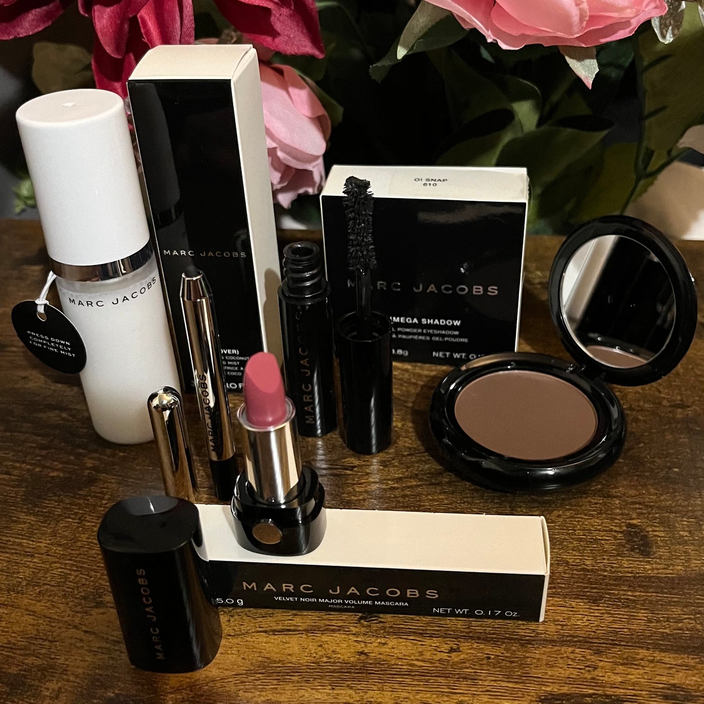 Elegant Marc Jacobs 6-piece luxury beauty gift collection featuring a glossy compact omega eyeshadow in the color “oh Boy”, A Limited Edition Signature Coconut Setting Spray, The Famous Award winning Highliner Gel Eye Crayon in the color “Blacquar” and an assortment of matching designer beauty cosmetics, complemented by a backdrop of lush pink, green & white florals.