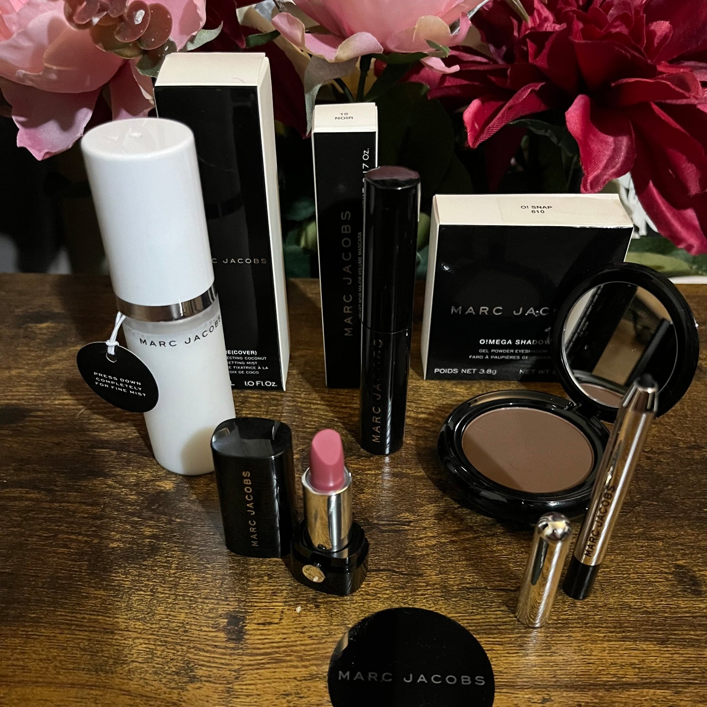 Elegant Marc Jacobs 6-piece luxury beauty gift collection featuring a glossy compact omega eyeshadow in the color “oh Boy”, A Limited Edition Signature Coconut Setting Spray, The Famous Award winning Highliner Gel Eye Crayon in the color “Blacquar” and an assortment of matching designer beauty cosmetics, complemented by a backdrop of lush pink, green & white florals.