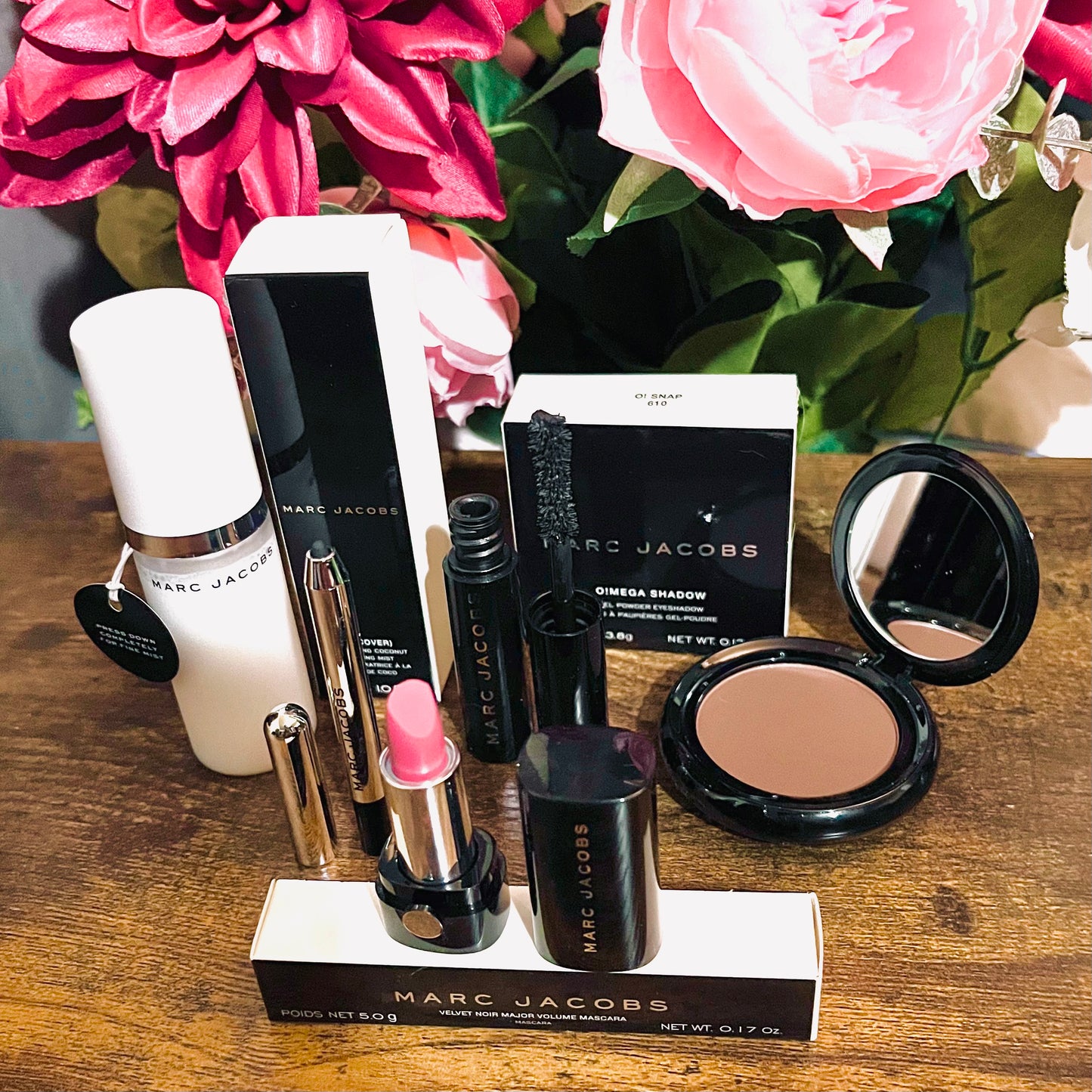 Elegant Marc Jacobs 6-piece luxury beauty gift collection featuring a glossy compact omega eyeshadow in the color “oh Boy”, A Limited Edition Signature Coconut Setting Spray, The Famous Award winning Highliner Gel Eye Crayon in the color “Blacquar” and an assortment of matching designer beauty cosmetics, complemented by a backdrop of lush pink, green & white florals.