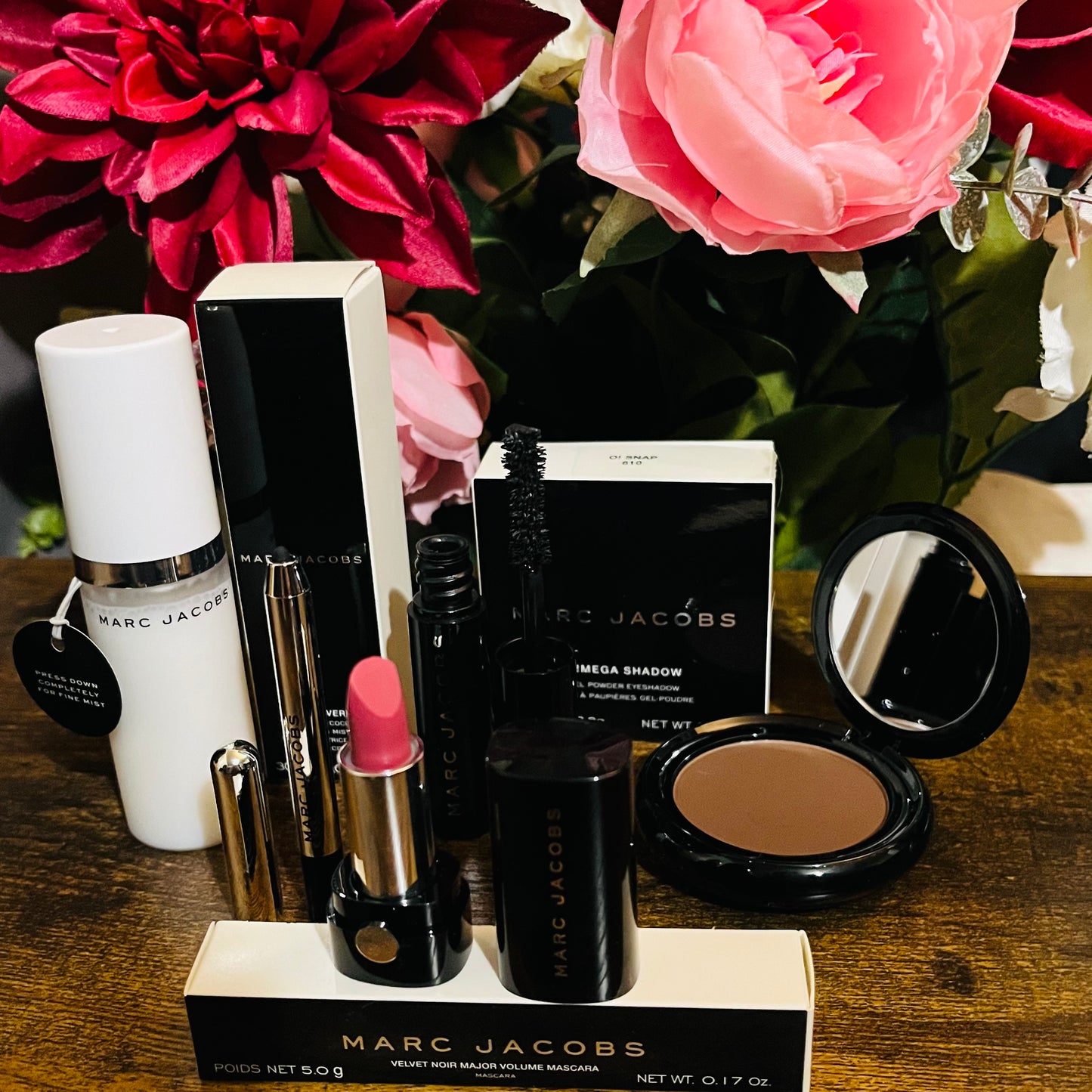 Elegant Marc Jacobs 6-piece luxury beauty gift collection featuring a glossy compact omega eyeshadow in the color “oh Boy”, A Limited Edition Signature Coconut Setting Spray, The Famous Award winning Highliner Gel Eye Crayon in the color “Blacquar” and an assortment of matching designer beauty cosmetics, complemented by a backdrop of lush pink, green & white florals.