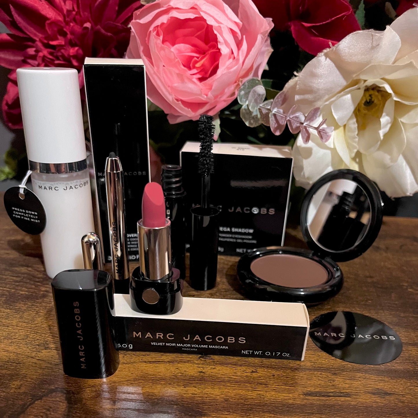 Elegant Marc Jacobs 6-piece luxury beauty gift collection featuring a glossy compact omega eyeshadow in the color “oh Boy”, A Limited Edition Signature Coconut Setting Spray, The Famous Award winning Highliner Gel Eye Crayon in the color “Blacquar” and an assortment of matching designer beauty cosmetics, complemented by a backdrop of lush pink, green & white florals.