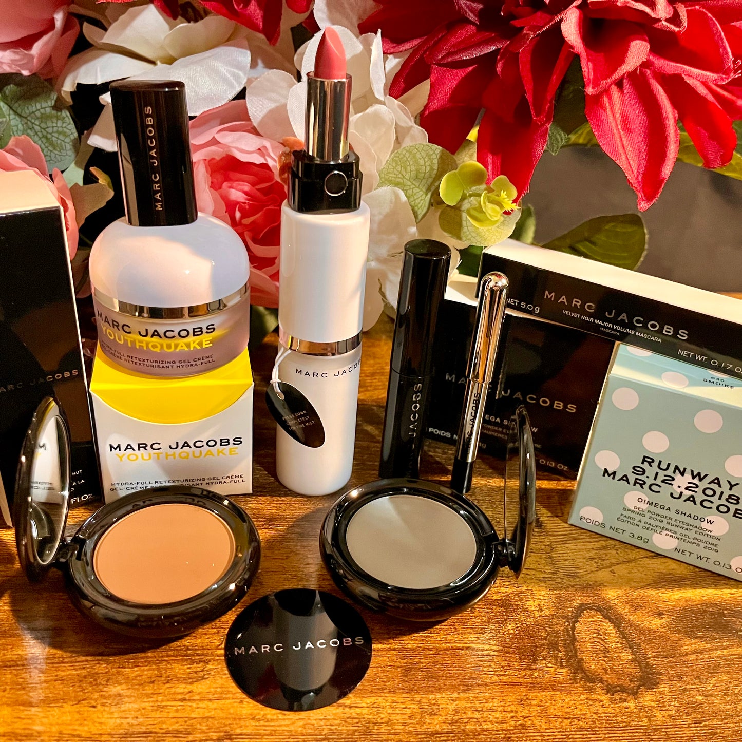 Elegant Marc Jacobs beauty collection featuring glossy compact powders, signature coconut setting spray, and assorted beauty cosmetics, complemented by a backdrop of lush pink & white florals.