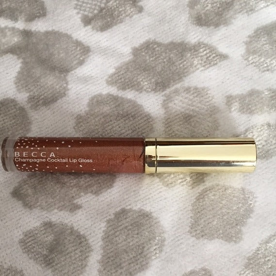 This Lip Gloss Is A very warm-toned, light-medium Tan (Soft Brown) with a hint of Peach pearl finish. It is a sheer lip-gloss that not sticky and goes on smooth and with ease. The 3D light-reflecting pigments as well as Ginger, Peppermint oil, and a non-stinging peptide make lips appear fuller and smoother. Contains Vitamins C and Vitamin E, plus hydrating Shea Butter and Muru Muru Butters to soften, protect and condition the lips. Becca Champagne Cocktail Lip Gloss shade "Bellini"