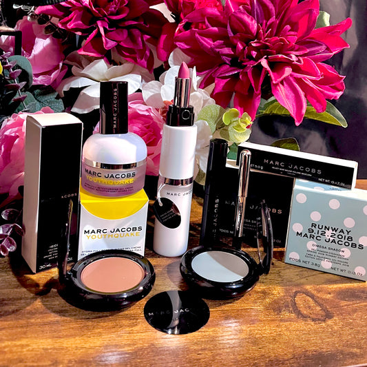 Elegant Marc Jacobs beauty collection featuring glossy compact powders, signature coconut setting spray, and assorted designer beauty cosmetics, complemented by a backdrop of lush pink & white florals.