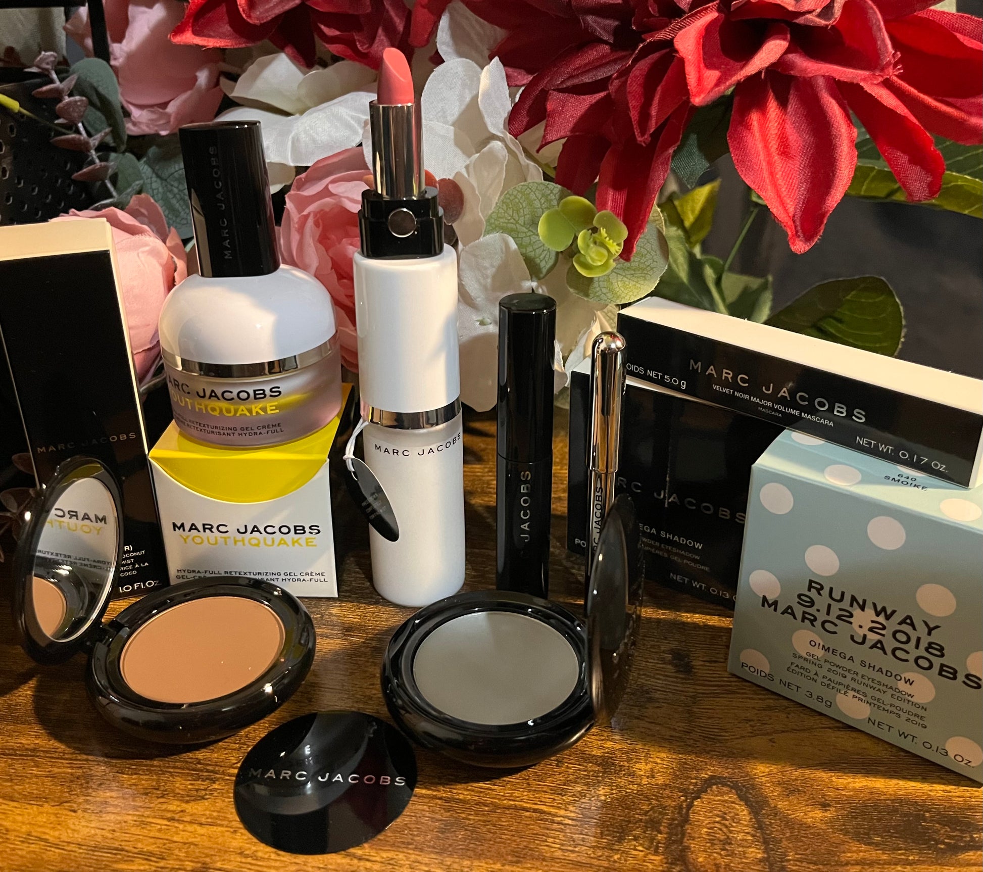 Elegant Marc Jacobs beauty collection featuring glossy compact powders, signature coconut setting spray, and assorted beauty cosmetics, complemented by a backdrop of lush pink & white florals.