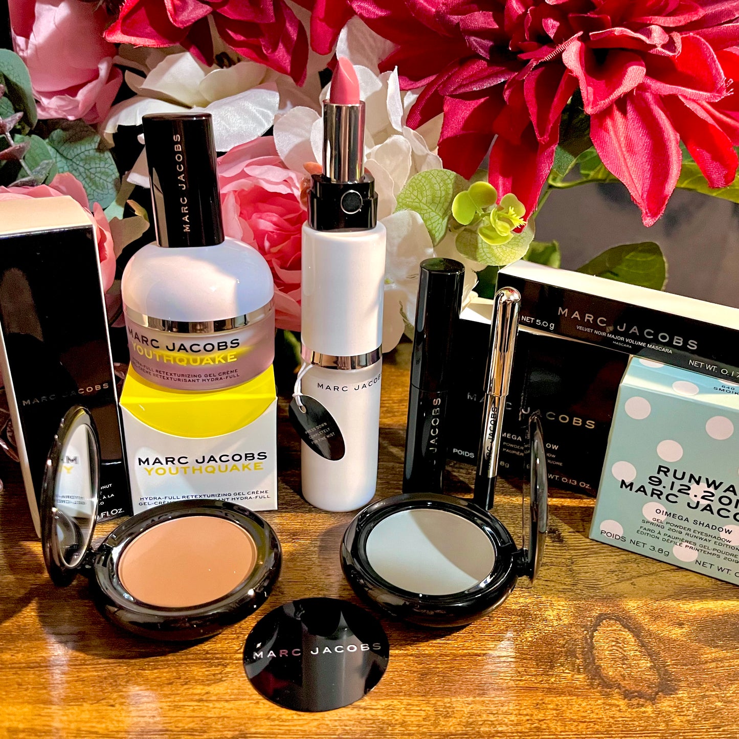 Elegant Marc Jacobs beauty collection featuring glossy compact powders, signature coconut setting spray, and assorted beauty cosmetics, complemented by a backdrop of lush pink & white florals.